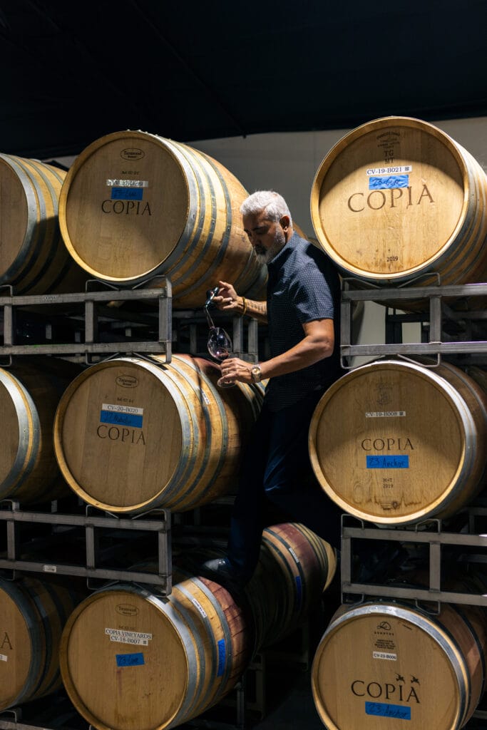 a man climbing in between barrels pouring wine into a glass from a wine thief